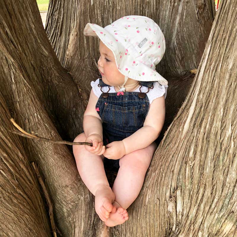 JAN & JUL Kids Cotton Floppy Hat Purple Daisy, white with pink daisies, on a baby sitting in a tree trunk.