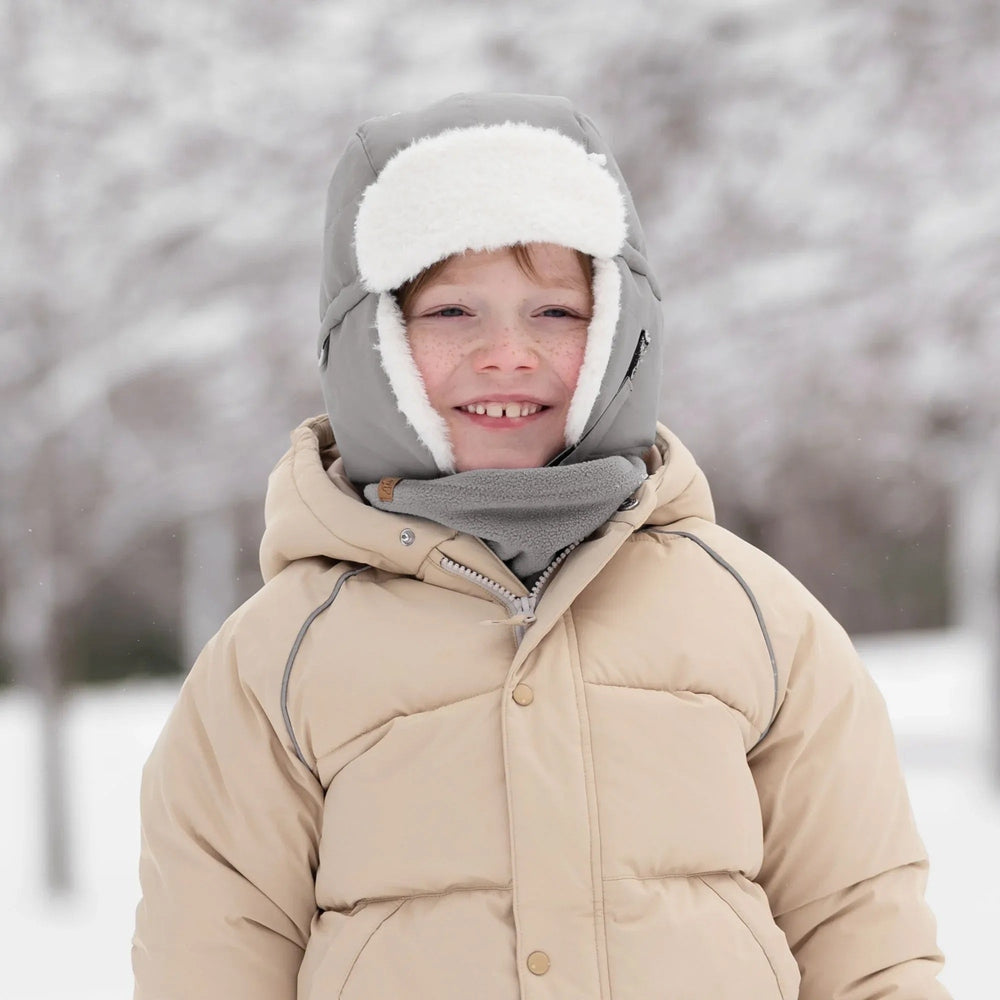 Jan & Jul Kids Insulated Winter Hats Greystone worn by a smiling child in a snowy park, highlighting gray fabric and fluffy white faux fur.