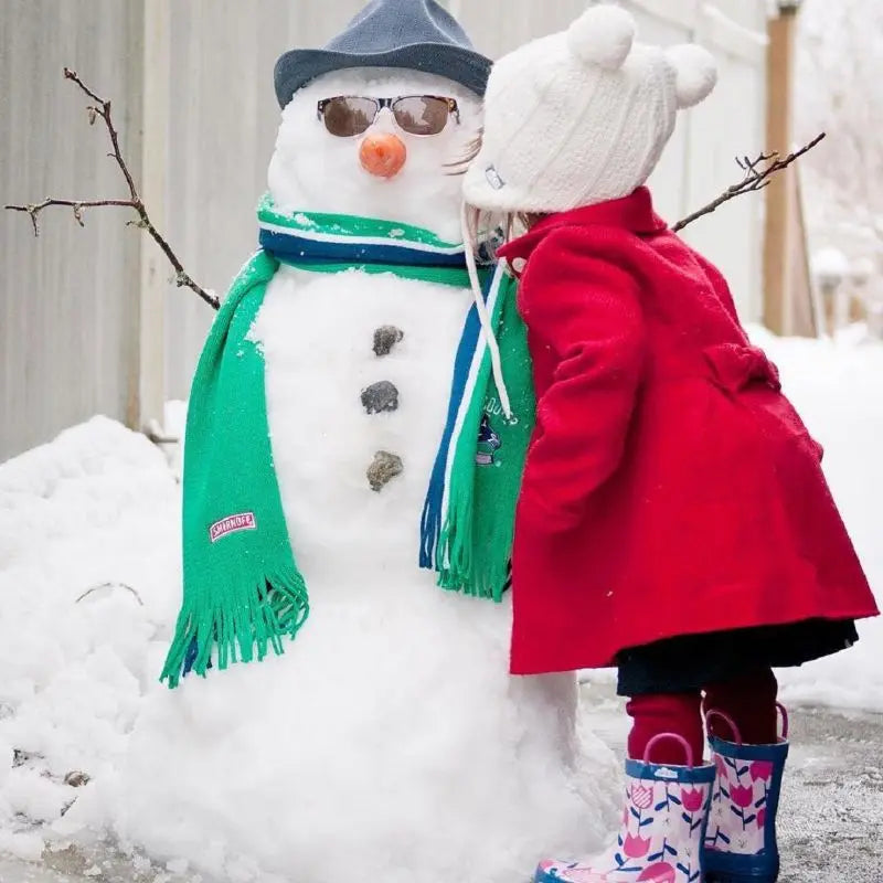 JAN & JUL Kids Knitted Beanies Hat Cream Bear worn by a child in a red coat kissing a snowman in a cold, wintry scene.