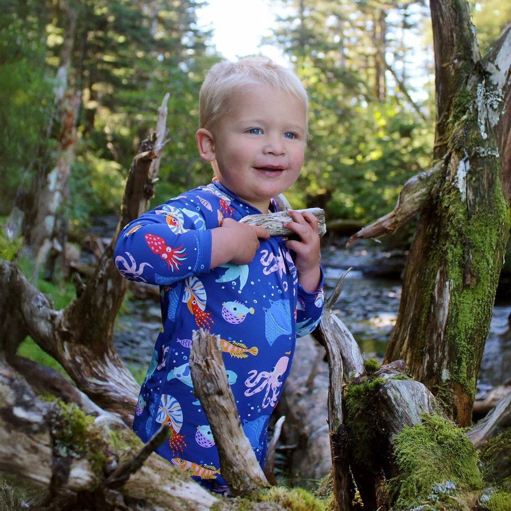 JAN & JUL Kids One Piece UV Sun Suit Sea Friends in blue aquatic print; toddler explores outdoors with a driftwood twig.