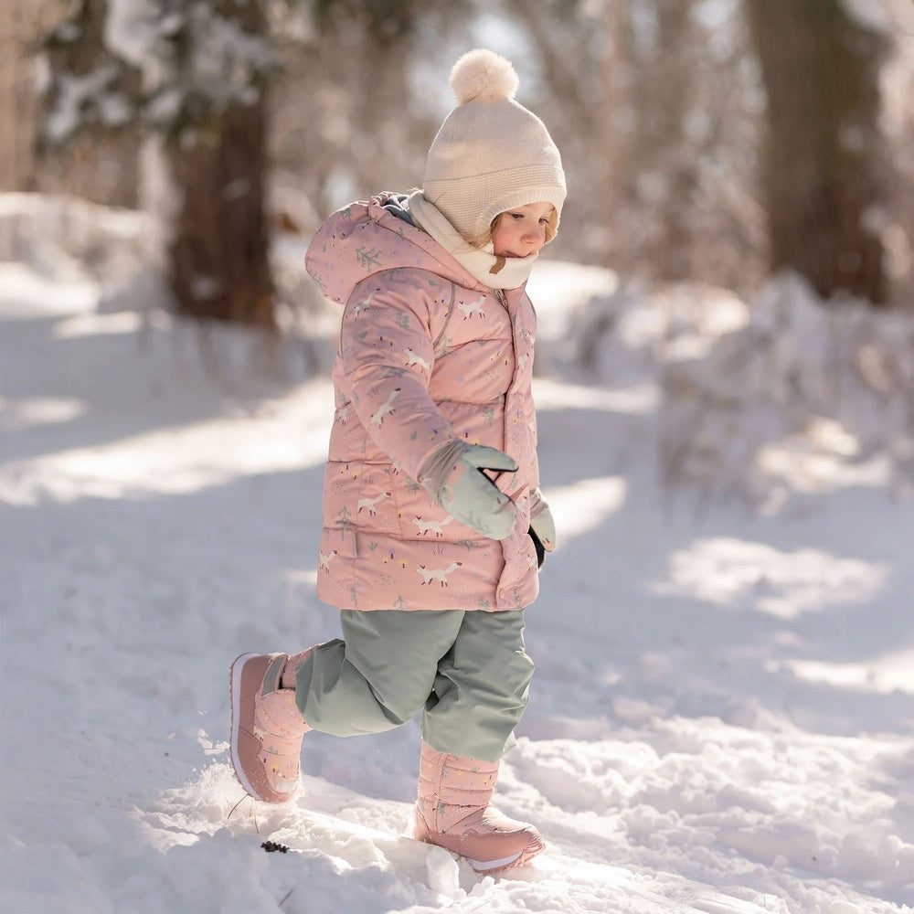 JAN & JUL Kids Pom-Pom Knit Hat Vanilla Cream worn by a toddler in a pink winter coat amid snowy trees.