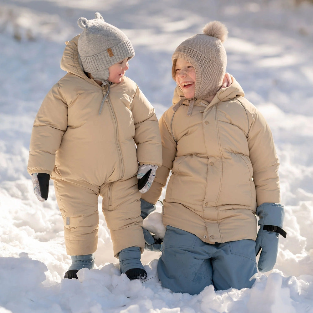 JAN & JUL Kids Pom-Pom Knit Hats Wheat Field in beige; two toddlers in matching winter snowsuits playing in the snow