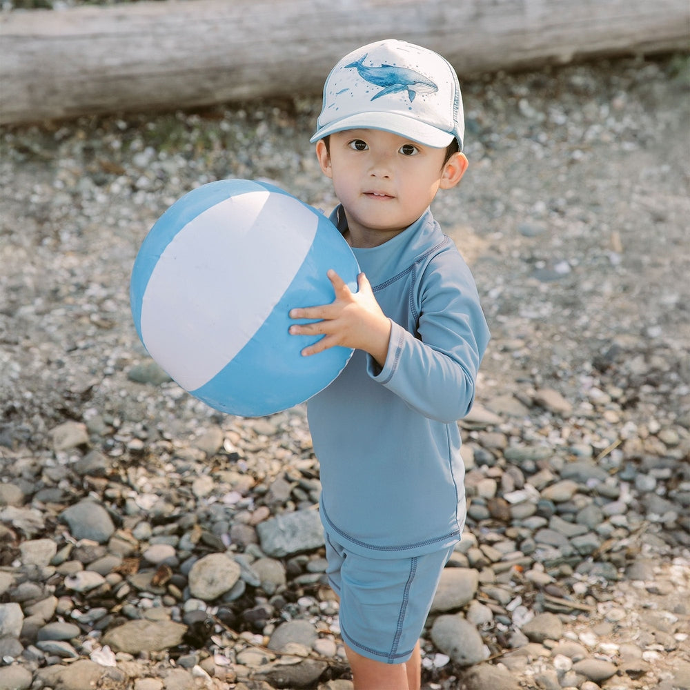Jan & Jul Kids UV Rashguard Top Blue Grey worn by toddler on a rocky beach, gripping a blue-white ball