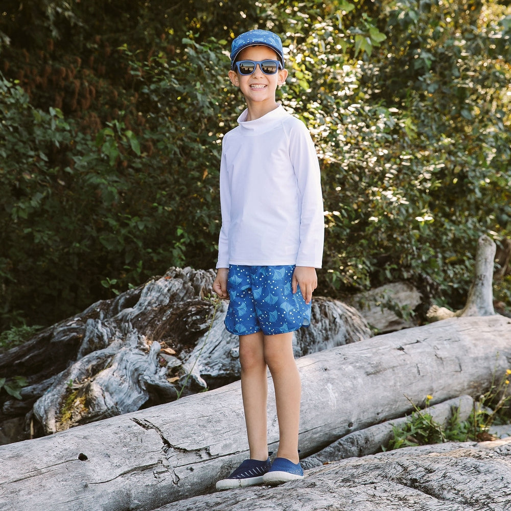 Jan & Jul Kids UV Swim Shorts Manta Ray in blue, worn by a smiling child standing on a log outdoors, showing the manta ray print.