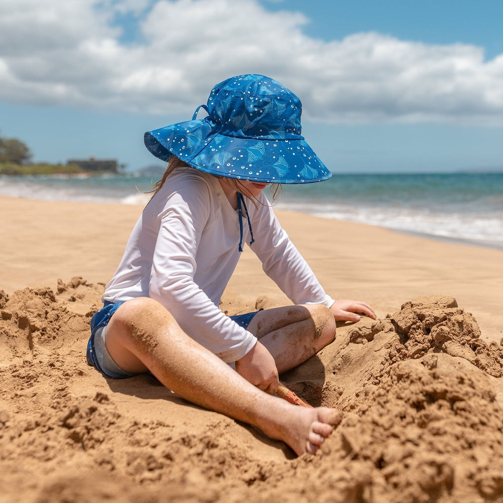 JAN & JUL Kids Water Repellent Adventure Hat Manta Ray blue wide-brim sunhat worn by a child on a sandy beach.