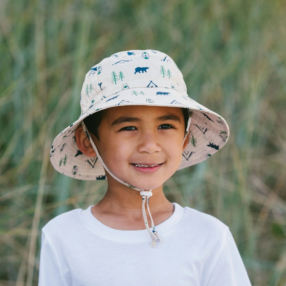 JAN & JUL Kids water-repellent Bear Camp bucket hat in white with teal camping icons, worn by smiling boy