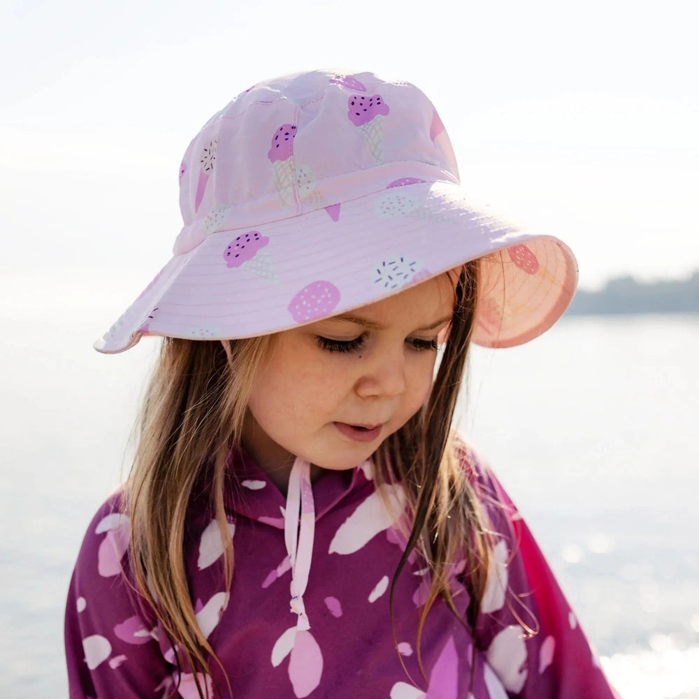 Jan & Jul Kids Water Repellent Bucket Hat Pink Ice Cream print worn by a girl by the sunny sea.