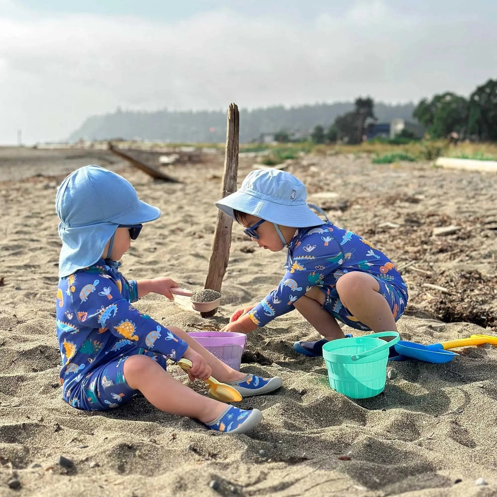 JAN & JUL Dino Buddies kids water shoes, blue dinosaur print, worn by two kids playing in the sand with buckets.