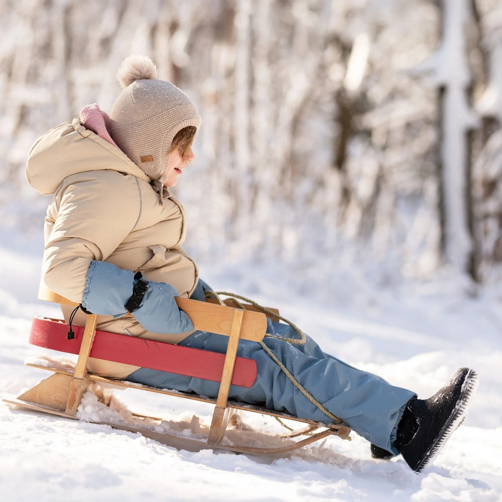 JAN & JUL Kids Waterproof Mittens Dusty Blue on a child sledding through a snowy forest.