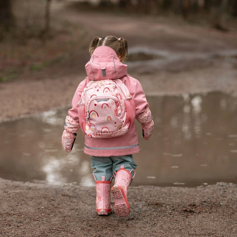 Jan & Jul Mini Backpacks pink rainbow pattern worn by a child walking along a muddy path with boots.