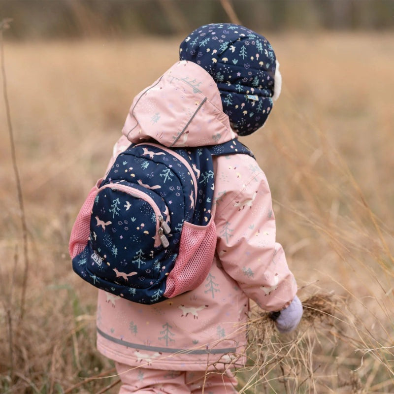JAN & JUL Mini Backpacks in navy/pink woodland print, worn by a child in a pink coat while gathering dried grass.