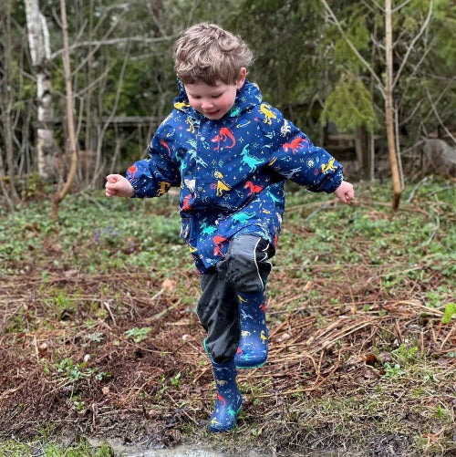 Jan & Jul Puddle Dry Rain Boots Space Dinos: toddler splashes in a muddy puddle wearing blue dinosaur-print boots.