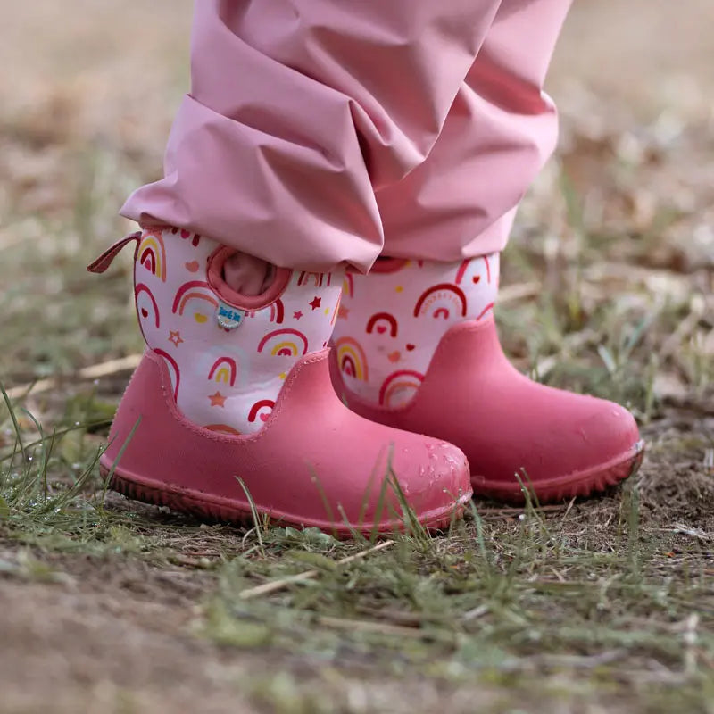 JAN & JUL Toasty Dry Lite Winter Boots Pink Rainbow; pink waterproof toddler boots with rainbow print, worn by a child walking on grass.