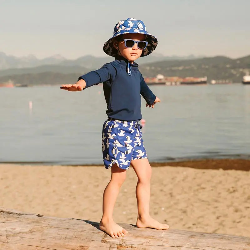 Jan & Jul Water Repellent Bucket Hats Seagull worn by a barefoot toddler walking along the beach.