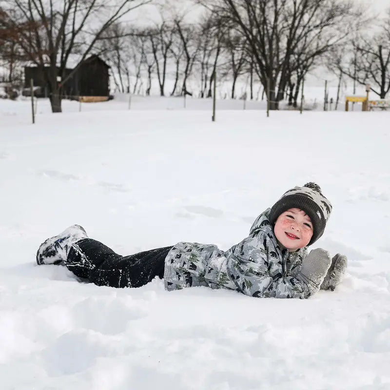 Jan & Jul Waterproof Cozy Dry Rain Jacket Bear for kids in gray camo, worn by a smiling child lying in the snow.