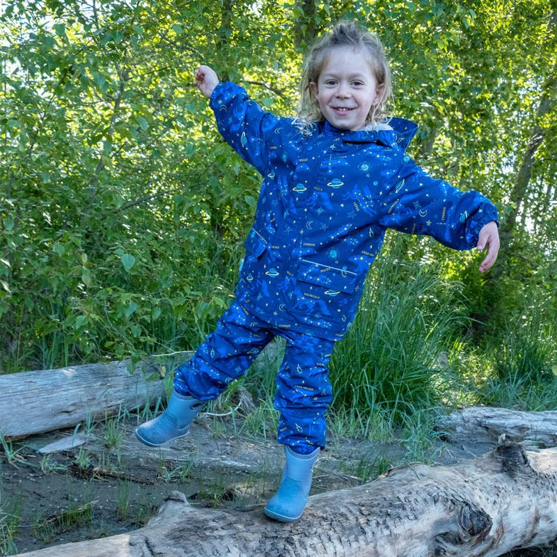 JAN & JUL Waterproof Cozy Dry Rain Pants Constellations worn by a smiling toddler balancing on a log in a blue constellation-print rain suit.