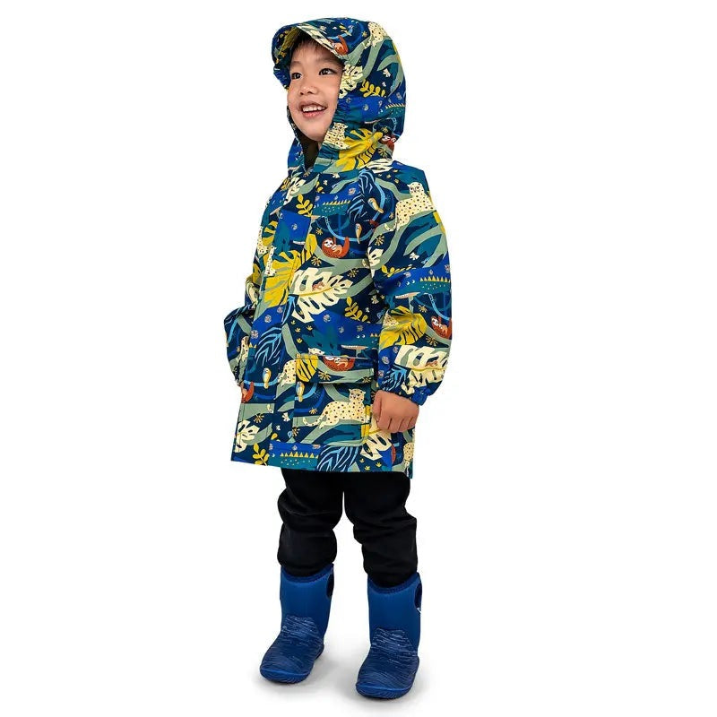 JAN & JUL Jungle Nights waterproof rain jacket with hood worn by a smiling kid in blue-yellow jungle print, blue rain boots.