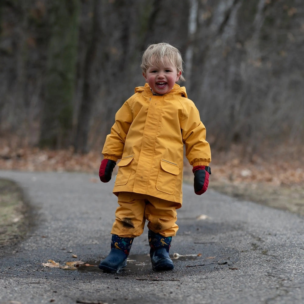 Jan & Jul Waterproof Puddle Dry Rain Jacket Yellow worn by a smiling toddler in a hooded coat with front pockets on a wet path.