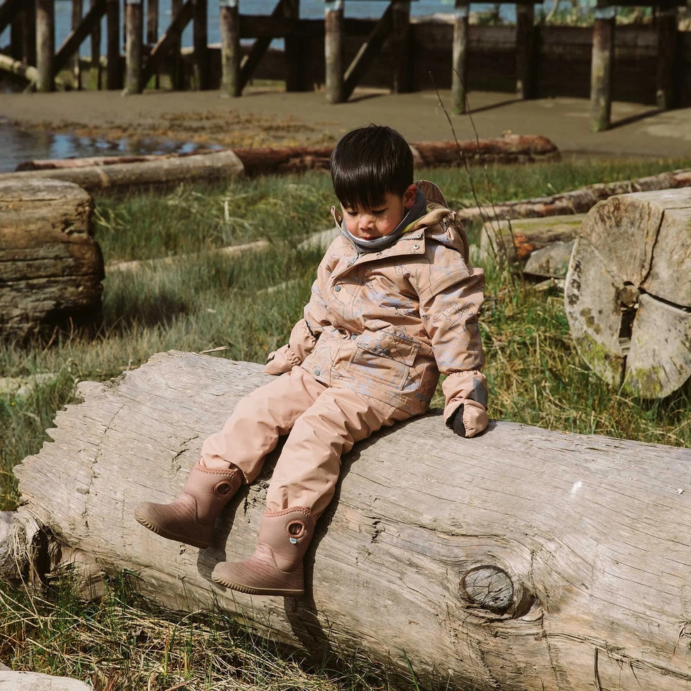 Toddler wearing Jan & Jul Waterproof Puddle Dry Rain Pants Soft Brown sits on a log outdoors, showcasing waterproof outerwear.