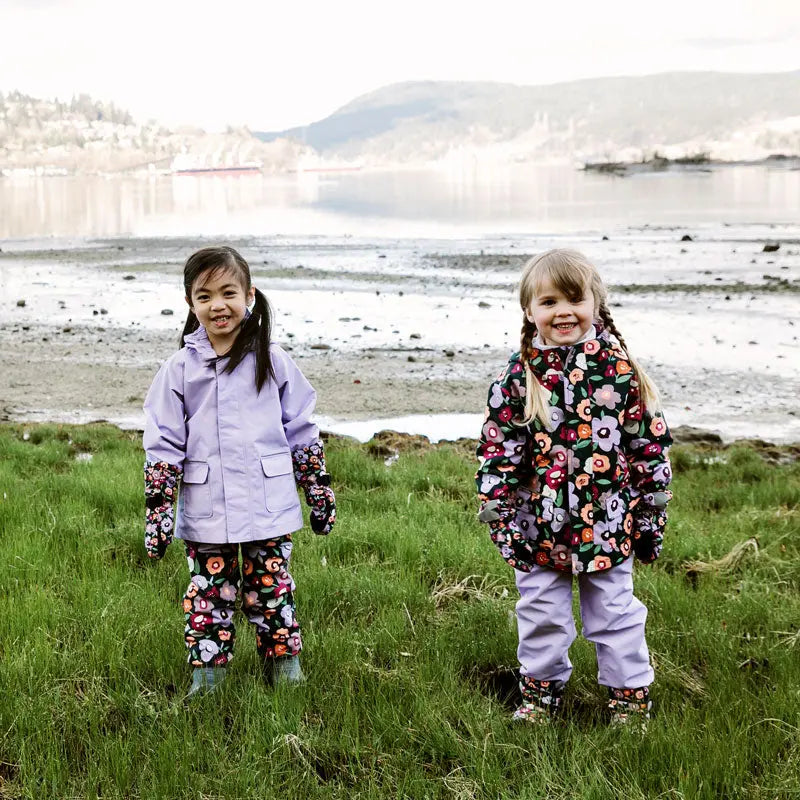 Jan & Jul Waterproof Puddle Dry Rain Pants with floral winter-flowers; two kids stand on grass by the shore in colorful jackets.