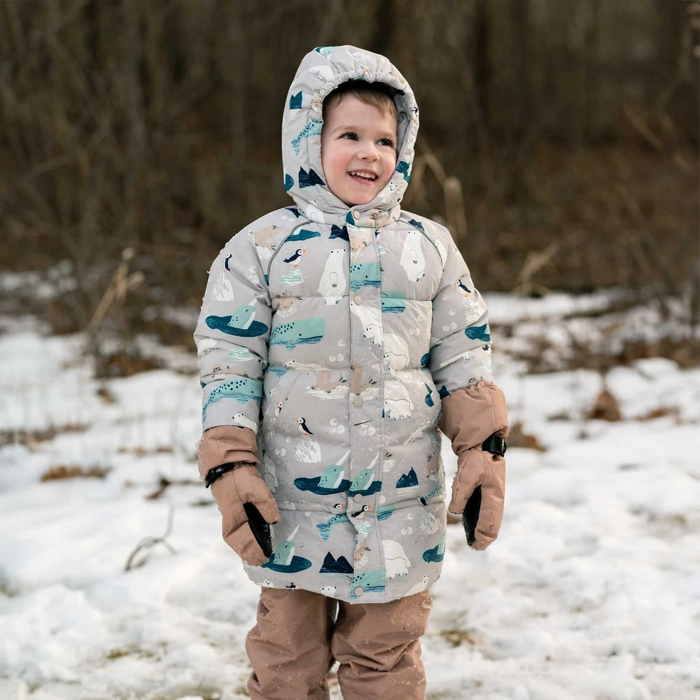 Jan & Jul Polar Pals waterproof winter coat on a smiling toddler in gray penguin print, hooded in snowy woods.