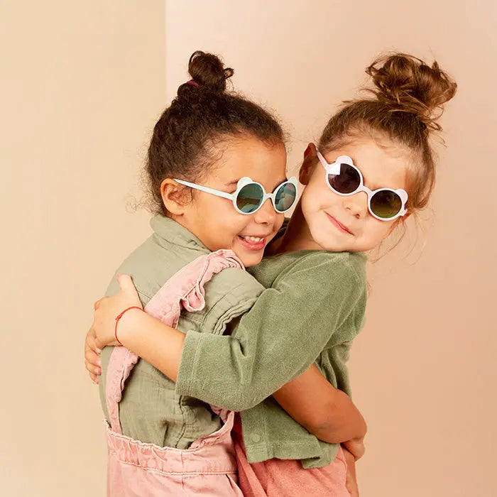 Two kids hug joyfully, wearing KI ET LA Sunglasses Ourson Light Pink with round white frames.