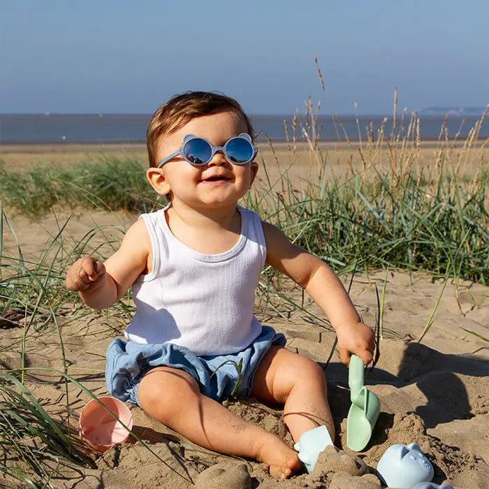 Ki Et La Sunglasses Ourson Silver Blue on a smiling baby at the beach, round blue lenses shielding eyes during play.