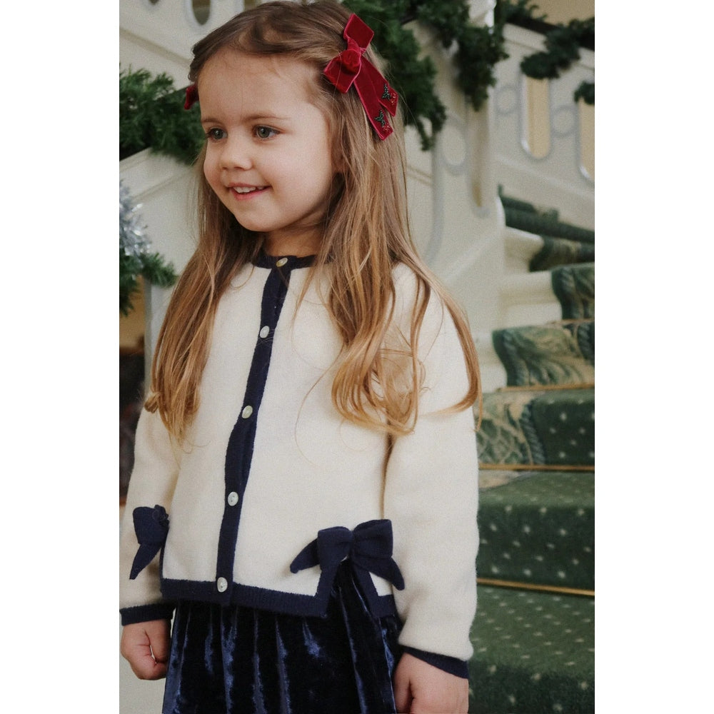 Konges Slojd Alette Cardigan Etoile in cream with navy trim and bows, worn by a smiling girl beside festive stairs.