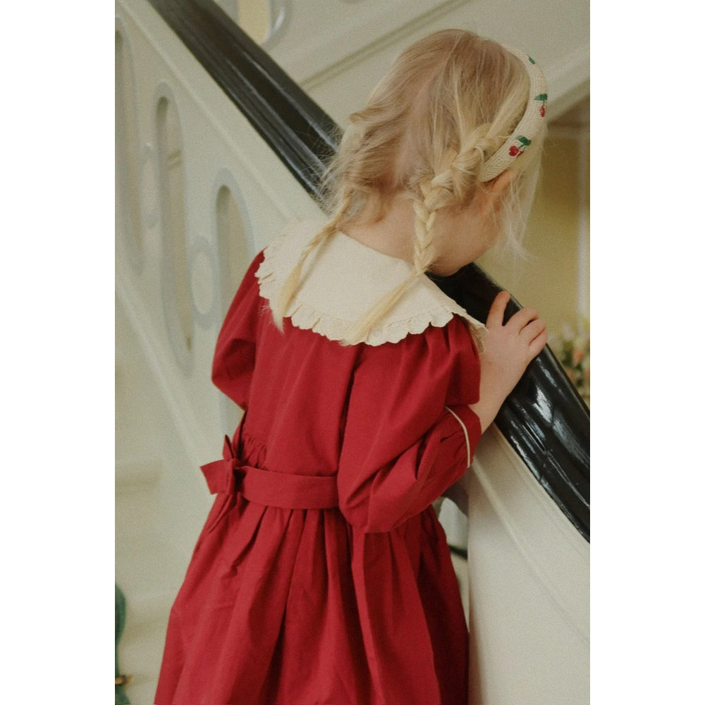 Konges Sløjd Coeur Dress Red Dahlia: toddler girl in red dress with white lace collar, braids, holding a railing.