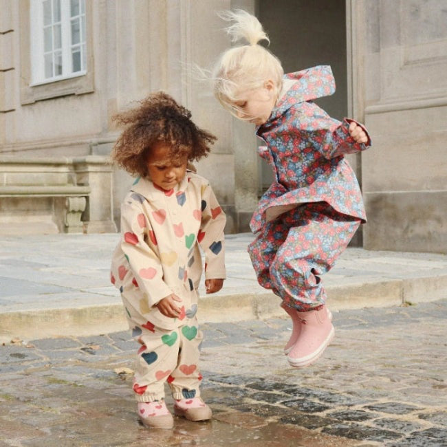Konges Slojd Faire Rain Set for toddlers; beige heart-print jumpsuit and blue floral raincoat, two kids jumping