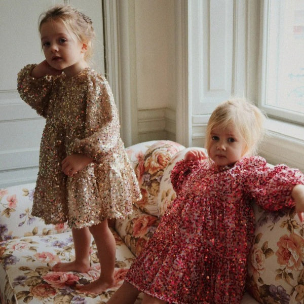 Konges Slojd Lila Sequins Bow Dress Gold Glitter; two toddlers wear sparkling sequin dresses on a floral sofa.