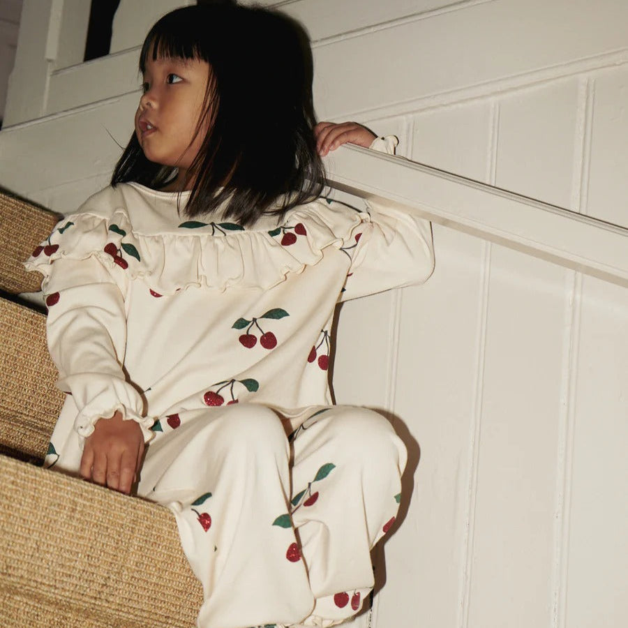 Konges Slojd Malli Glitter Blouse worn by a girl on stairs; cream top with cherry print and ruffled collar.