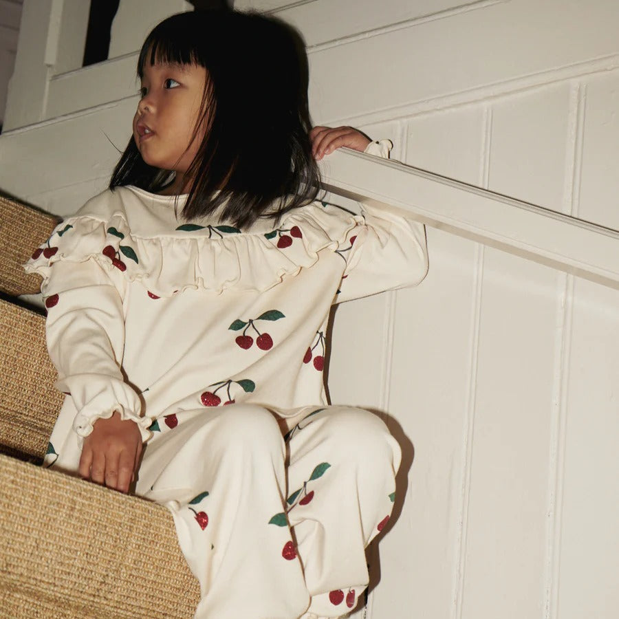 Konges Slojd Malli Glitter Pants worn by a child sitting on a staircase in cream cherry-print fabric.