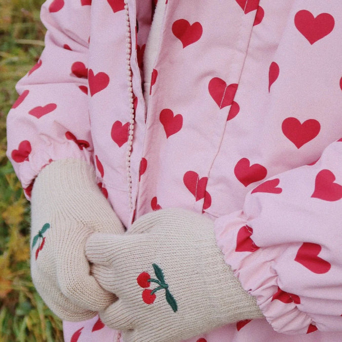 Konges Slojd Mismou Jacket Amour Pink worn by a child, heart-print outerwear with cream gloves embroidered with cherries.