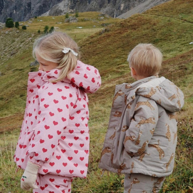 Konges Slojd Mismou Jacket Amour Pink: girl in pink heart-print coat and boy in tiger-print gray jacket on a cool hillside.