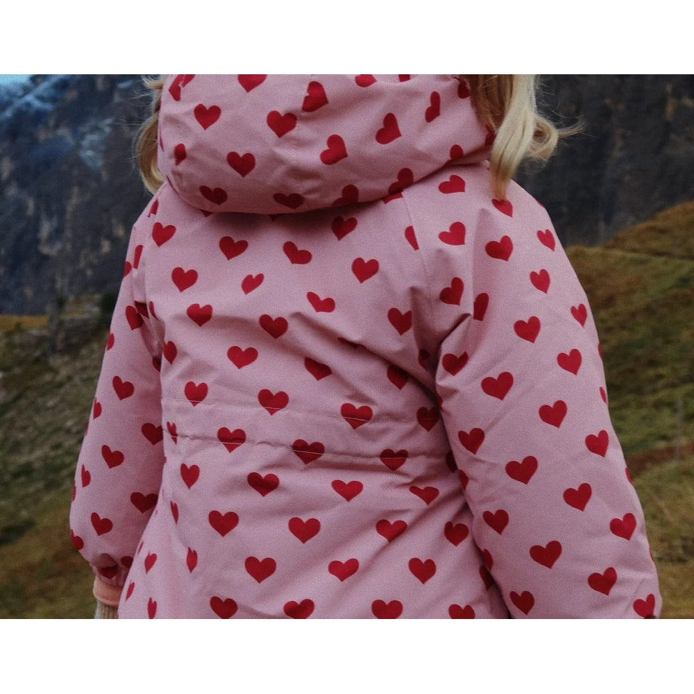 Konges Slojd Mismou Jacket Amour Pink worn by a child walking in a rocky outdoor landscape in a pink heart-patterned puffer with hood.