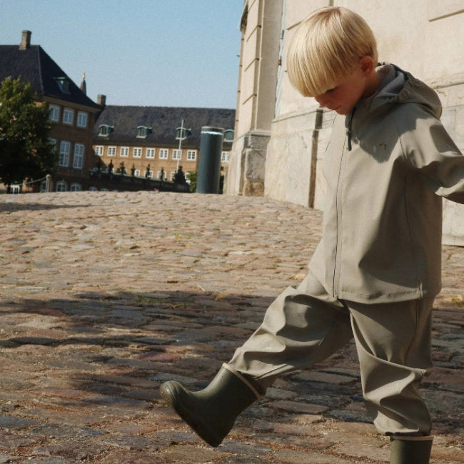 Konges Slojd Plainy Rain Set Laurel Oak toddler rain jacket and pants with olive boots on cobblestone street