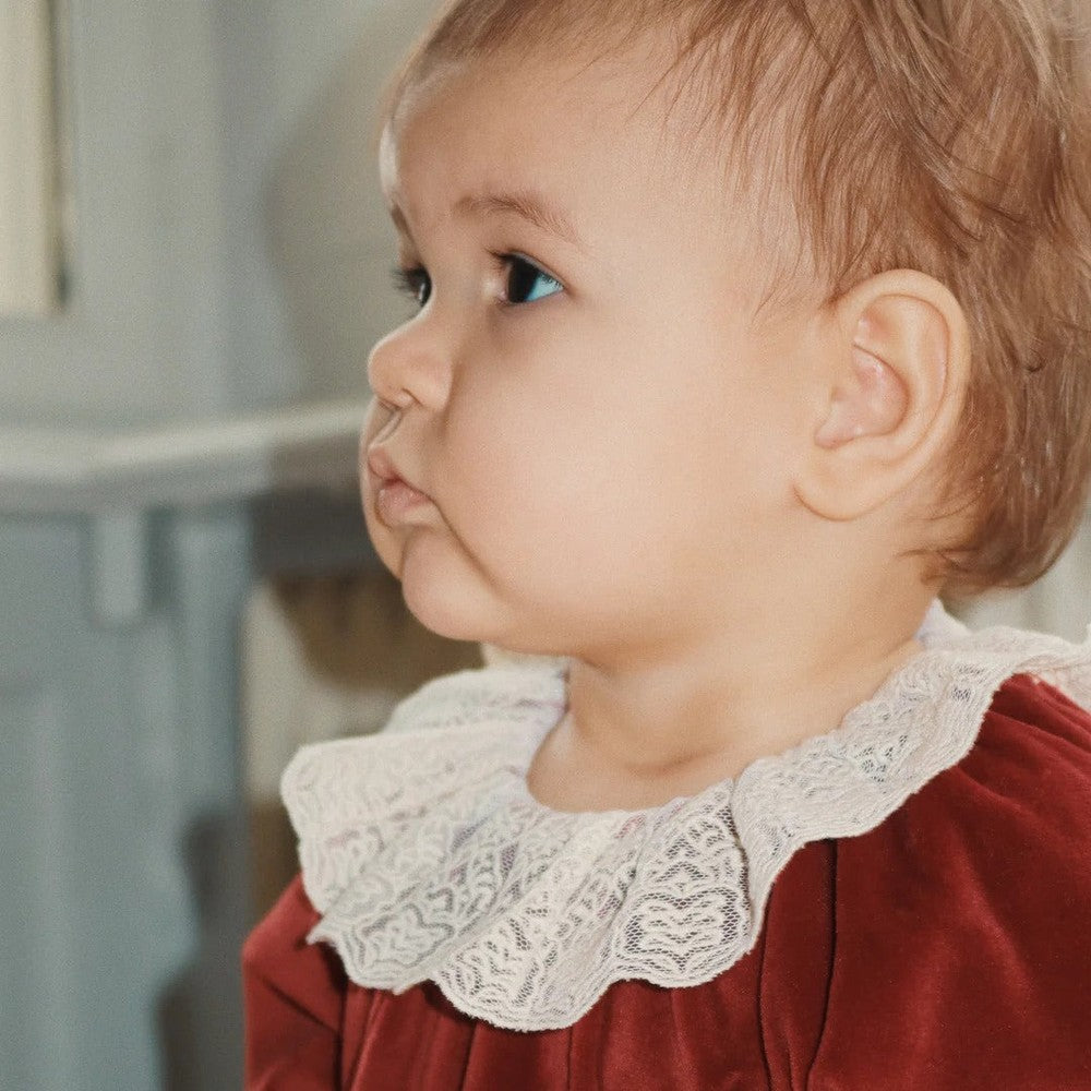 Konges Slojd Venola Romper Red Dahlia on a baby, plush red velvet one-piece with delicate white lace collar.
