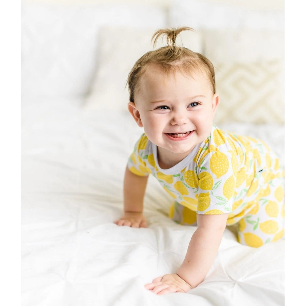 Little Sleepies Bamboo Pajama Set Lemons; a baby crawls on a white bed in lemon-print yellow pajamas.