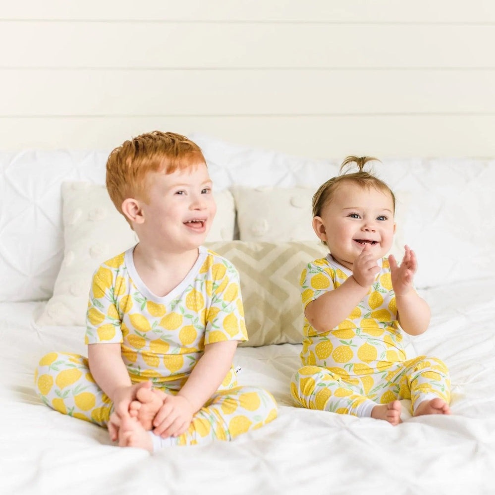 Little Sleepies Bamboo Pajama Set Lemons; two kids in matching yellow lemon-print bamboo pajamas on a white bed.