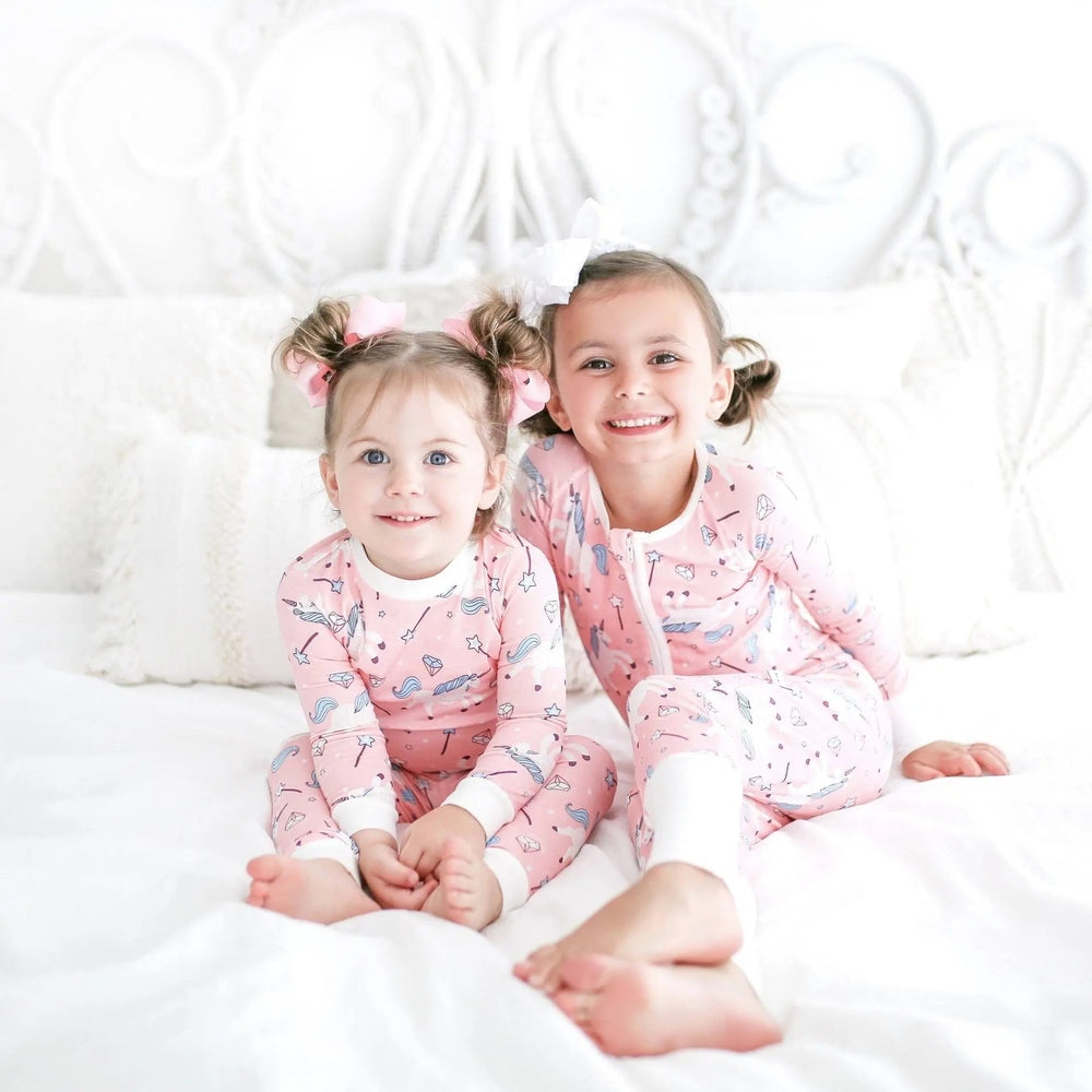 Little Sleepies Bamboo Pajama Set Pink Unicorn; two kids in matching pink unicorn pajamas sit and smile on a white bed.