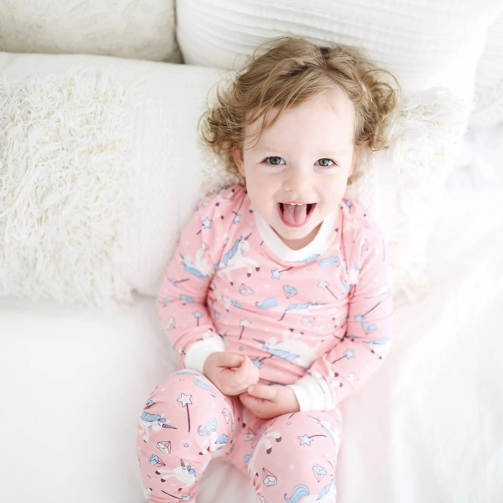 Little Sleepies Bamboo Pajama Set Pink Unicorn, toddler sits on a white sofa smiling in pastel unicorn-print sleepwear