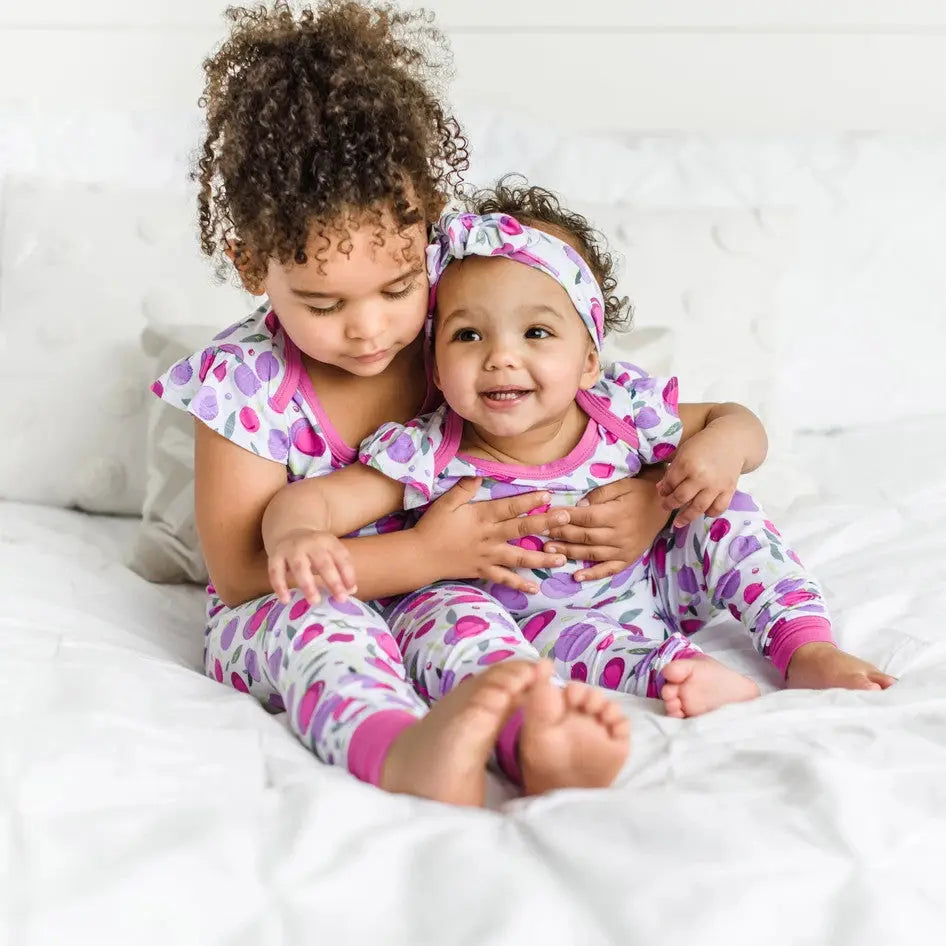 Little Sleepies Bamboo Pajama Set Plum: two siblings cuddle in matching plum-print PJs on a white bed.