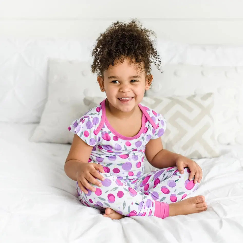 Little Sleepies Bamboo Pajama Set Plum worn by a smiling toddler sitting cross-legged on a white bed with pink-trim plum print.