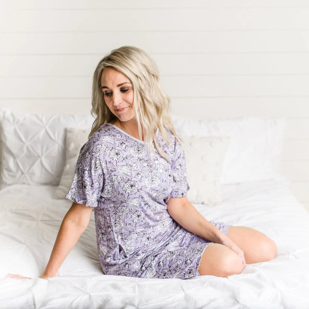 Little Sleepies Bamboo Pajama Set Women's Peonies in lavender, woman sits on a white bed in matching short-sleeve top and shorts.