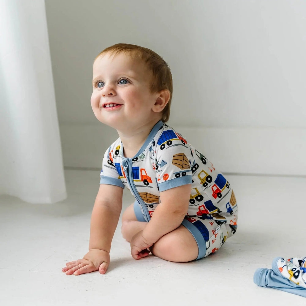 Littlesleepies Construction Bamboo Viscose Shorty Zippy romper in blue trim; baby crawls smiling at construction trucks.