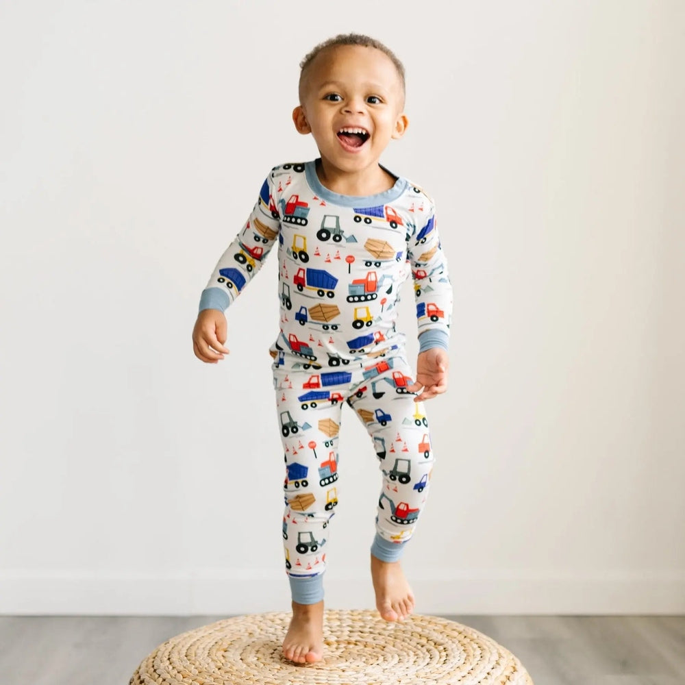 Littlesleepies Construction Two-Piece Bamboo Viscose Pajama Set; toddler jumps on rug in white print with construction vehicles and blue cuffs