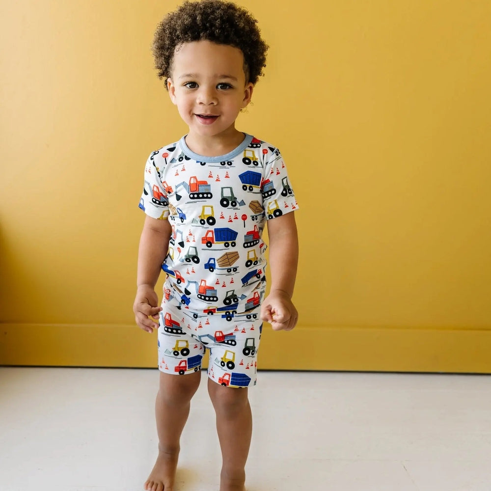 Littlesleepies Construction two-piece short-sleeve bamboo viscose pajama set; toddler stands smiling in truck-print pajamas.