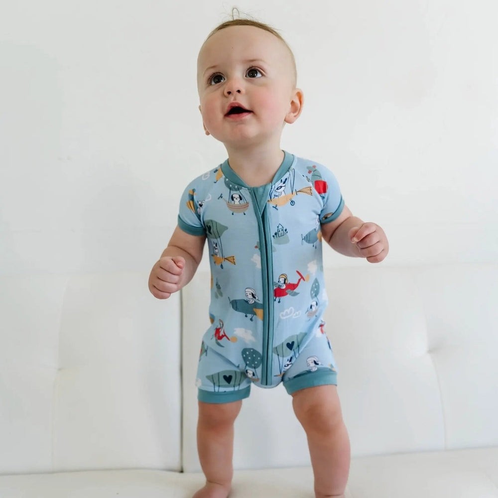 Littlesleepies Fly Away With Me bamboo viscose shorty zippy romper; baby stands on a white sofa in blue airplane print.