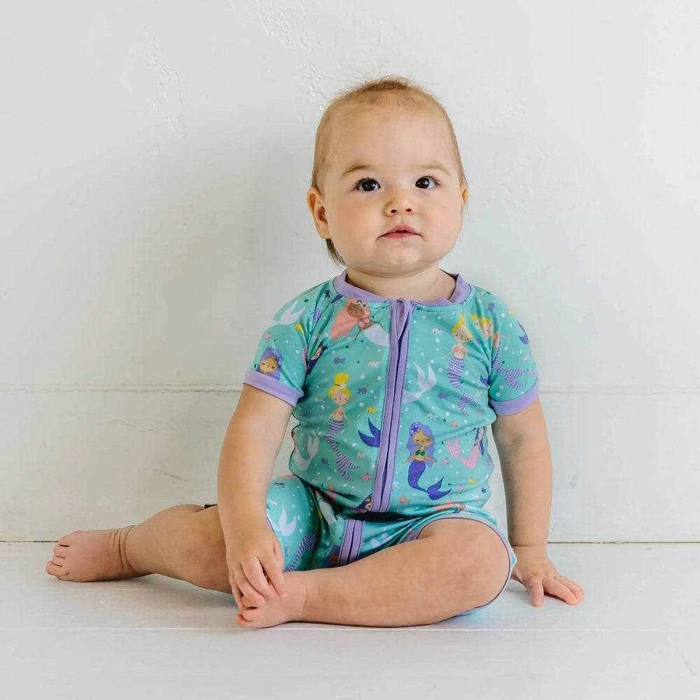 LITTLESLEEPIES Mermaid Magic Bamboo Viscose Shorty Zippy romper on a baby sitting cross-legged in teal with purple trim and a front zipper.